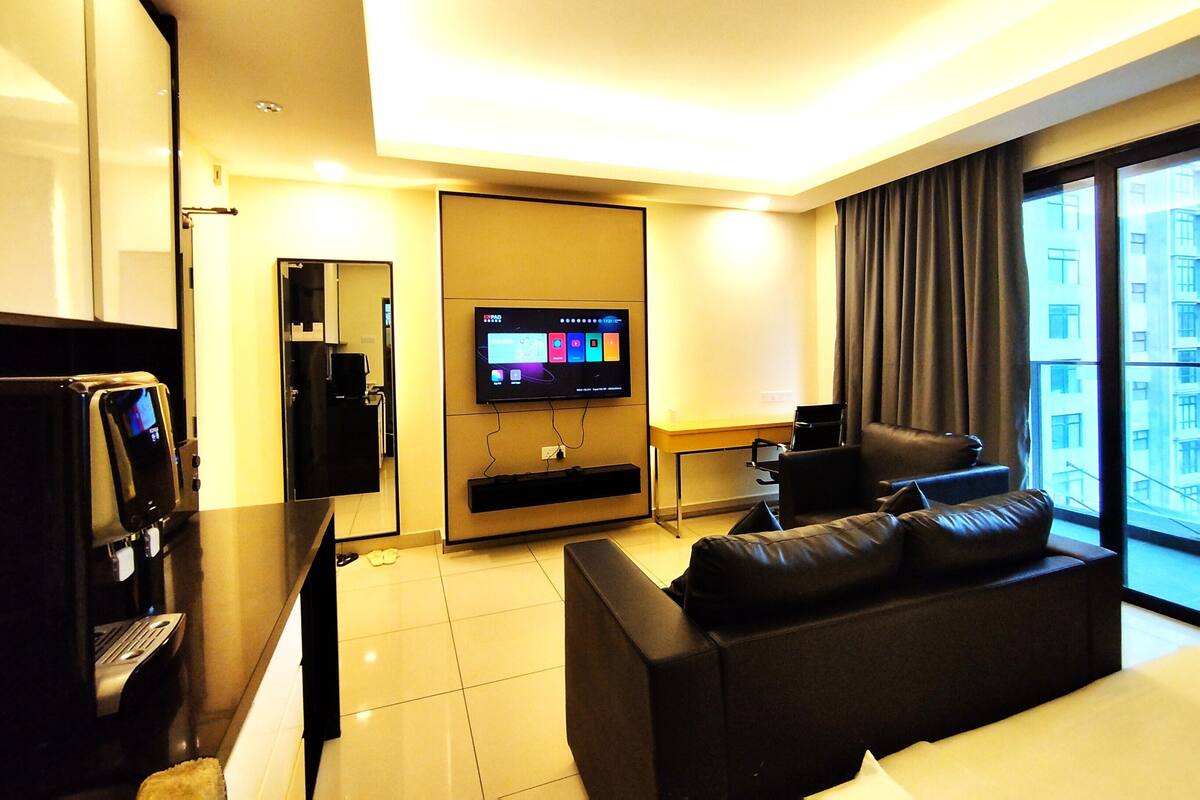 A modern hotel room with a flat-screen TV mounted on the wall, a leather sofa, a small table with a coffee machine, and a large window offering a view of the city.