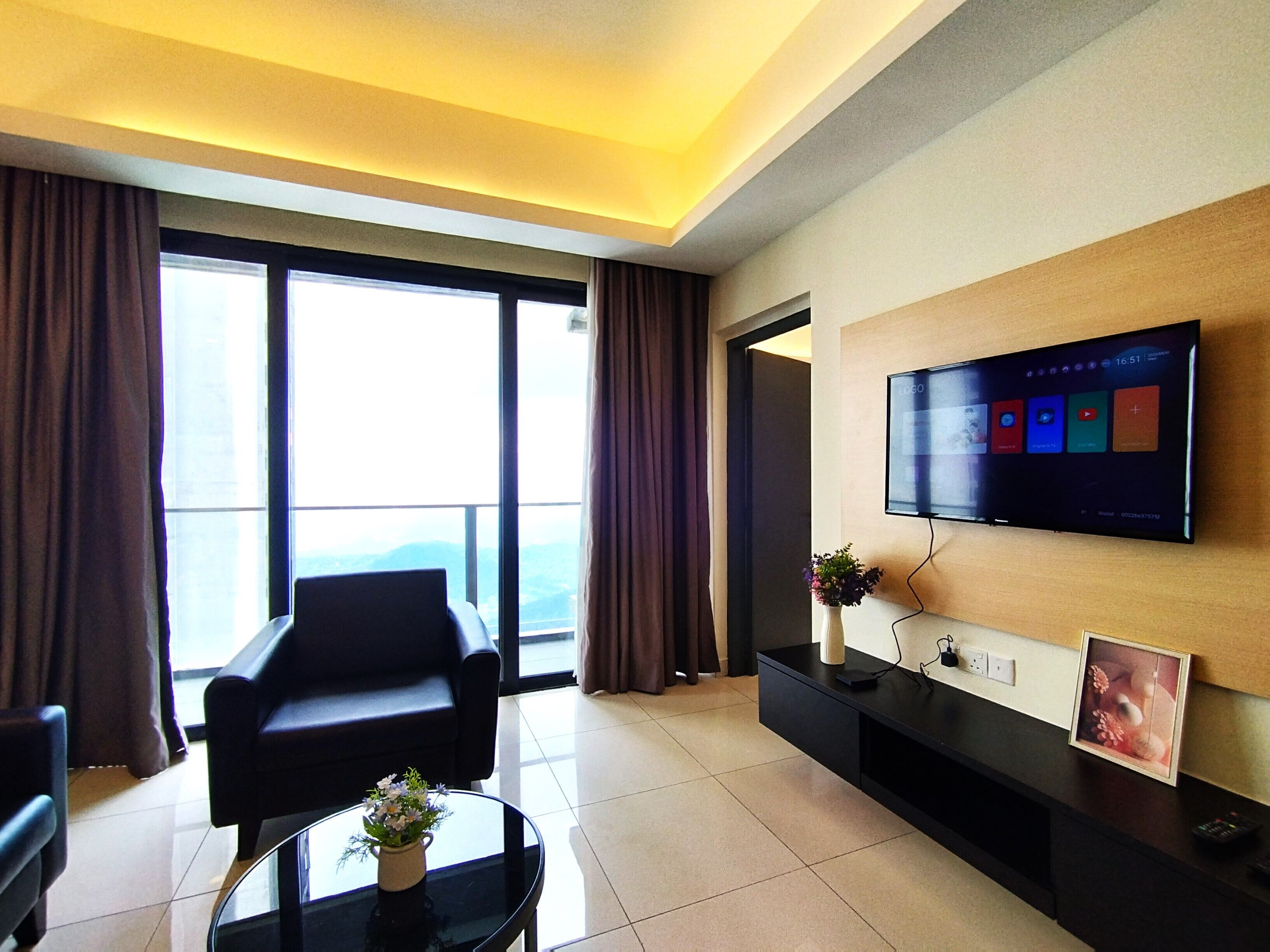 A modern hotel room with a large window, a flat-screen TV mounted on the wall, a black leather sofa, a small round table with a vase of flowers, and a black console table with a picture frame and remote controls.
