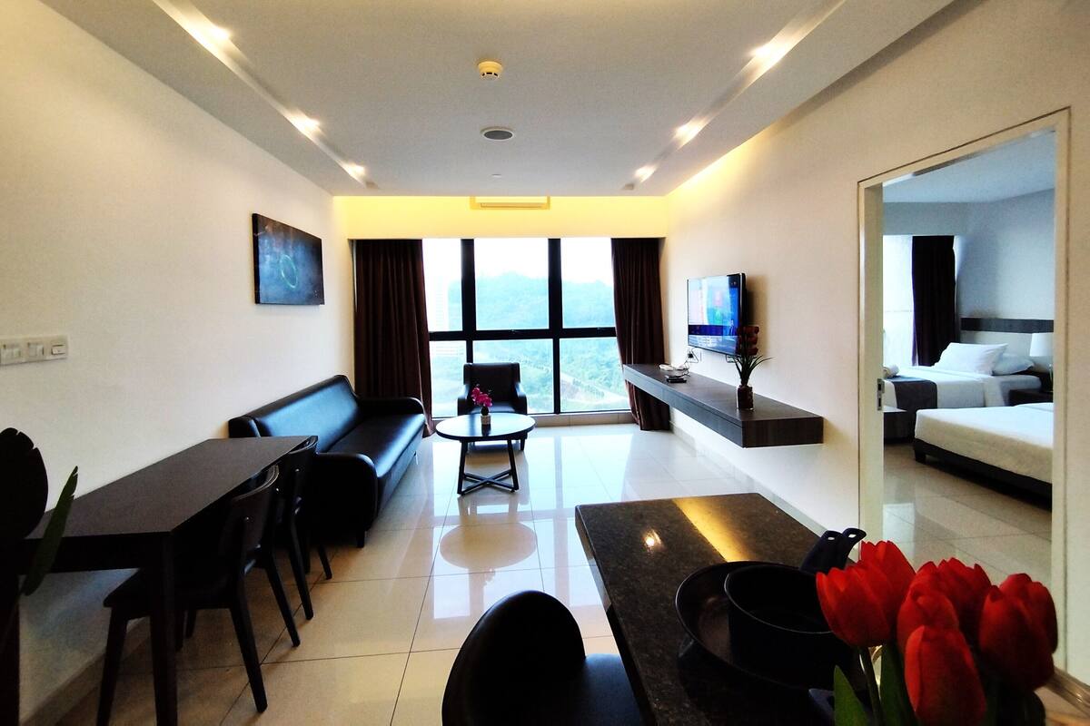 A modern hotel room with a sofa, dining table, chairs, a TV, and a large window offering a city view.