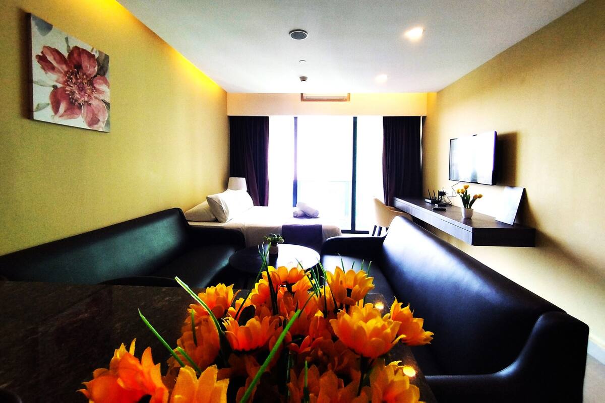 A hotel room with a sofa, a small table with a vase of flowers, a bed, a TV, and a window with curtains.