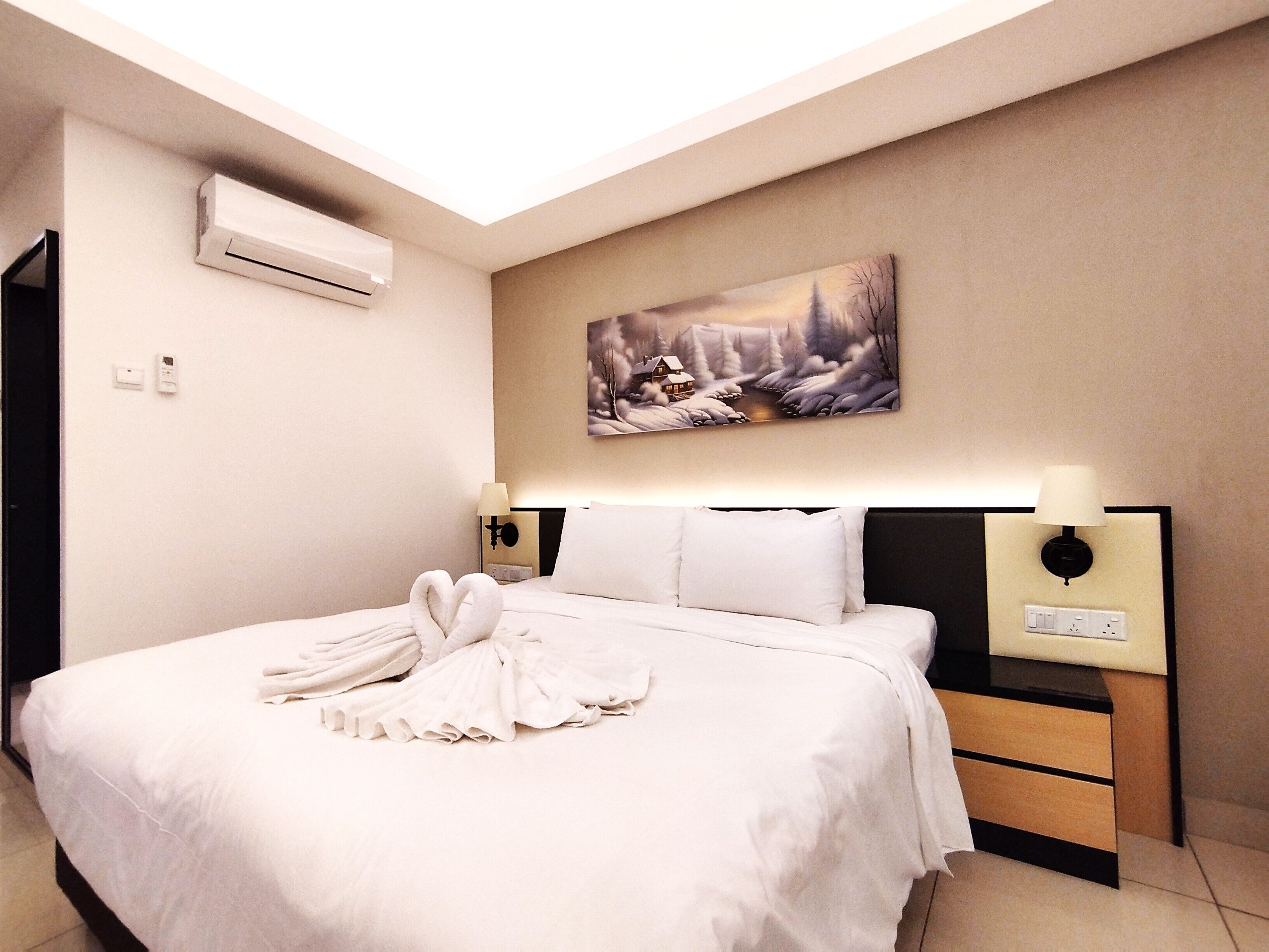 A neatly made bed with a heart-shaped towel arrangement, a painting of a winter scene, and a modern bedside table with a lamp.