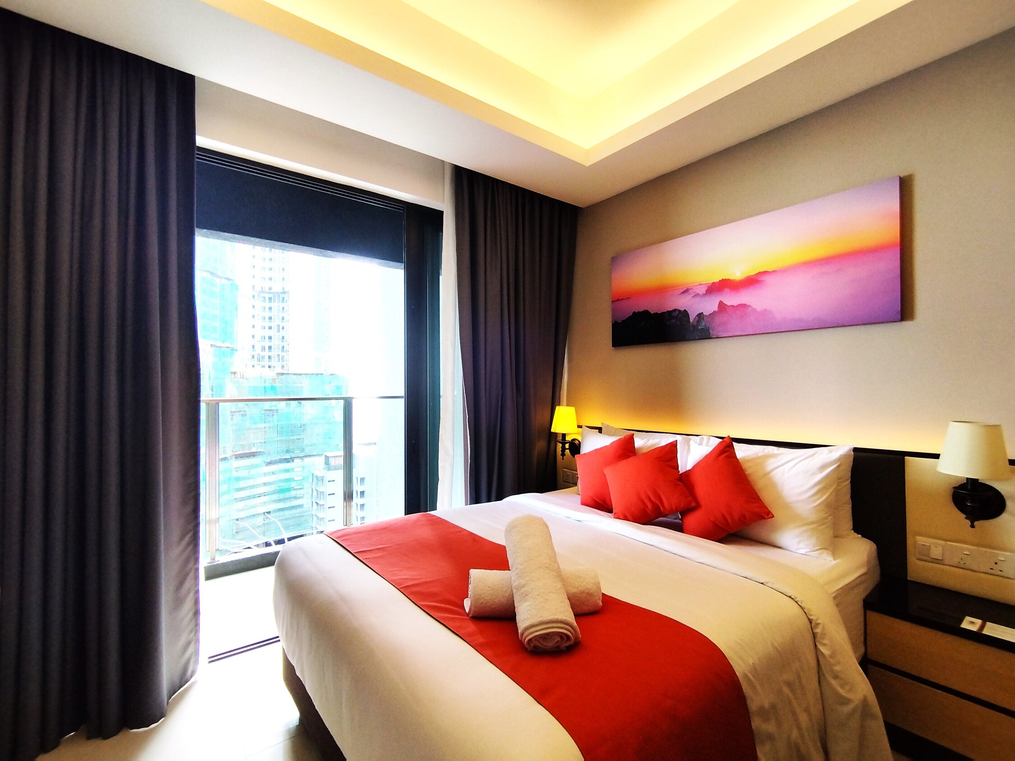 Een hotelkamer met een groot bed, rood-witte beddengoed, een schilderij van een zonsondergang, een nachtkastje met een lamp en uitzicht op gebouwen door het raam.