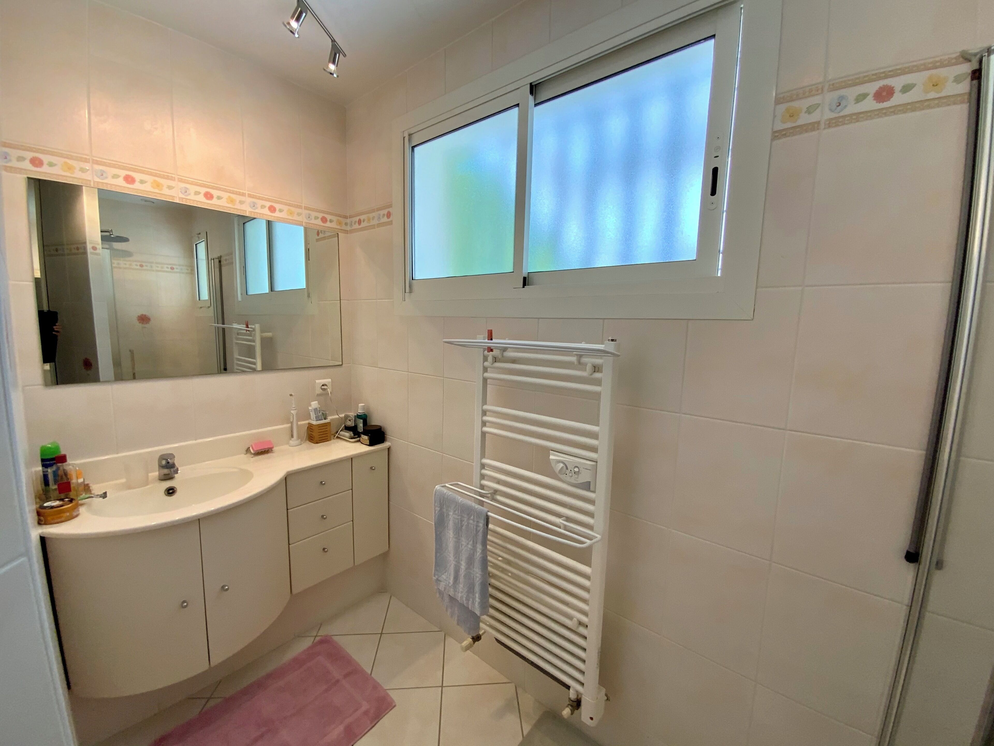 Ein modernes Badezimmer mit einem großen Spiegel, einem weißen Waschbecken, einem Handtuchhalter und einem Fenster über der Dusche.