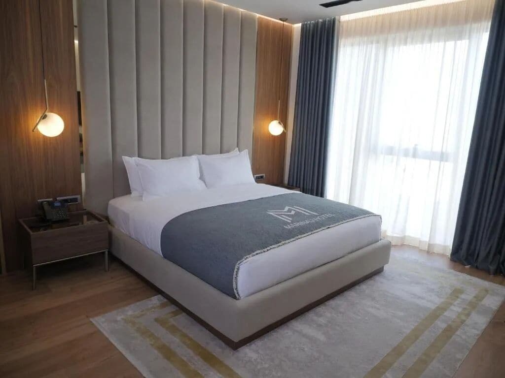 A modern hotel room with a large bed, a bedside table with a phone, and a view of the outdoors through large windows.