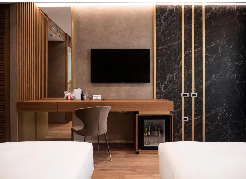 Una habitación de hotel moderna con un escritorio de madera, un televisor de pantalla plana montado en la pared, un minibar y una zona de estar con dos camas blancas.