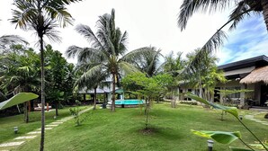 A well-maintained garden with palm trees, a pool, and a thatched-roof building.
