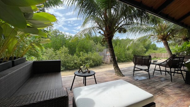 An outdoor patio with wicker furniture, a small table with a vase, and a view of a lush garden with palm trees.