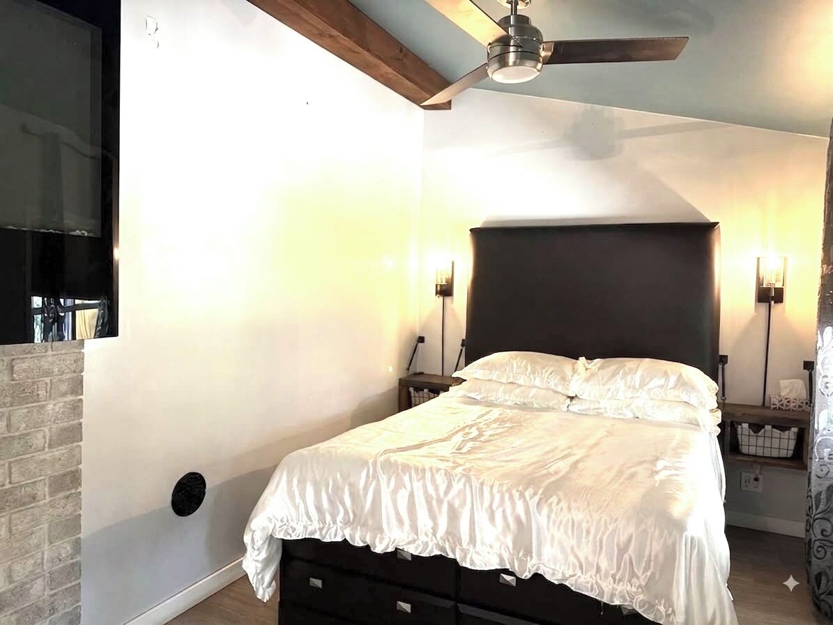 Un dormitorio con una cama, un ventilador de techo, dos lámparas de pared, una pared de ladrillo y una viga de madera en el techo.