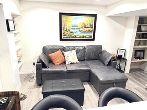 A modern living room with a gray sectional sofa, a small ottoman, and a framed landscape painting on the wall.