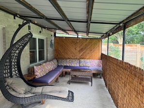 Un espacio al aire libre techado con una silla colgante, bancos acolchados y una mesa de mimbre.