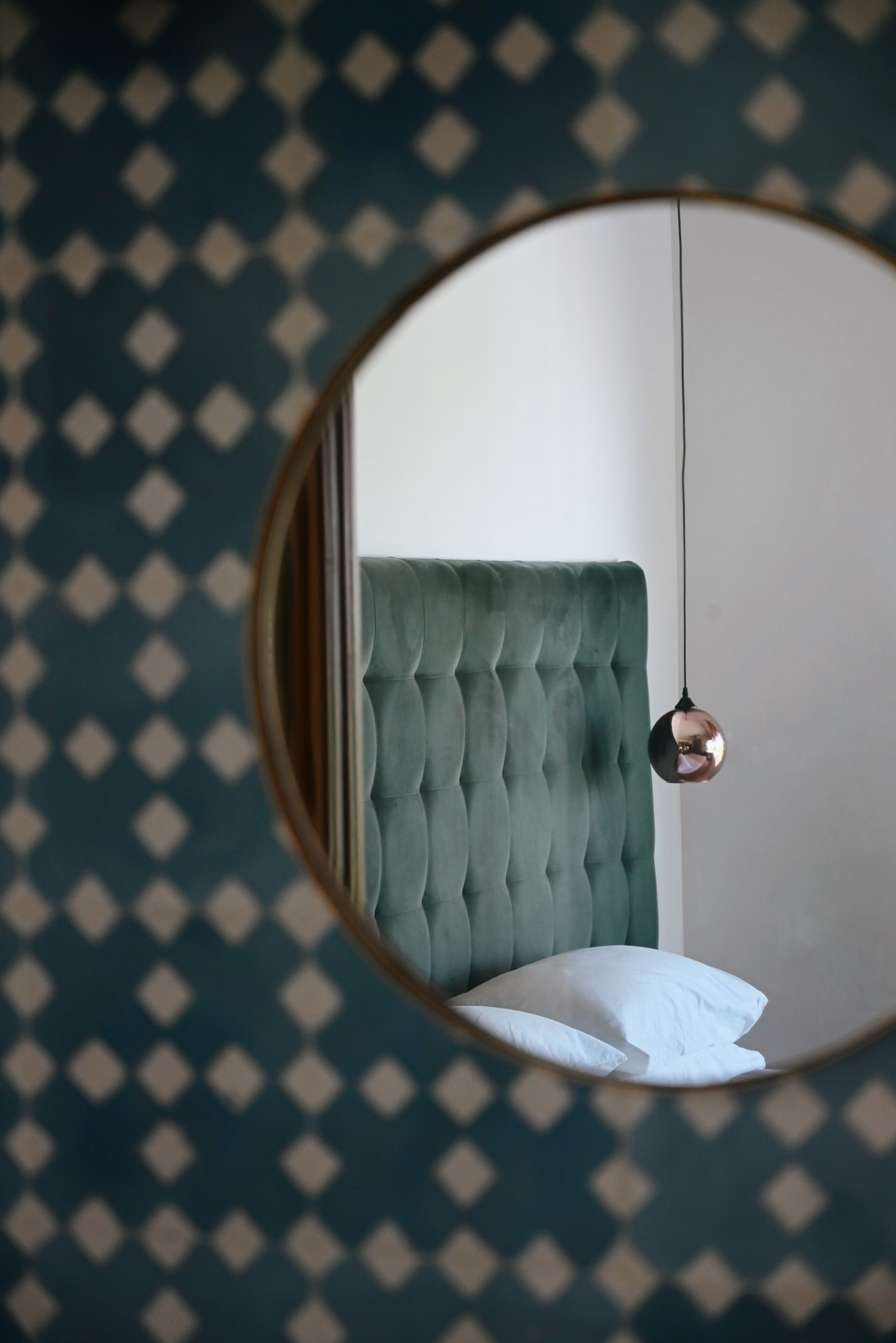 Espejo redondo que refleja una cama con cabecera verde y una lámpara colgante.