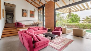 A spacious living area with a large red sofa set, a coffee table, and a view of a pool and greenery. - Muza Beach Villa Steps to Most Requested Beach! (Puntarenas Province)