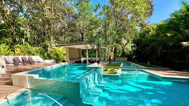 Une piscine rectangulaire à l’eau d’un bleu cristallin, entourée d’une végétation luxuriante et dotée d’un espace détente abrité avec des chaises longues.