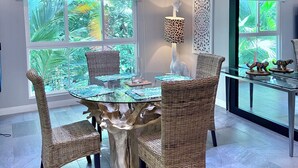 Un coin repas avec une table à plateau de verre entourée de chaises en osier, un luminaire décoratif suspendu et une vue sur une végétation luxuriante grâce à de grandes fenêtres.