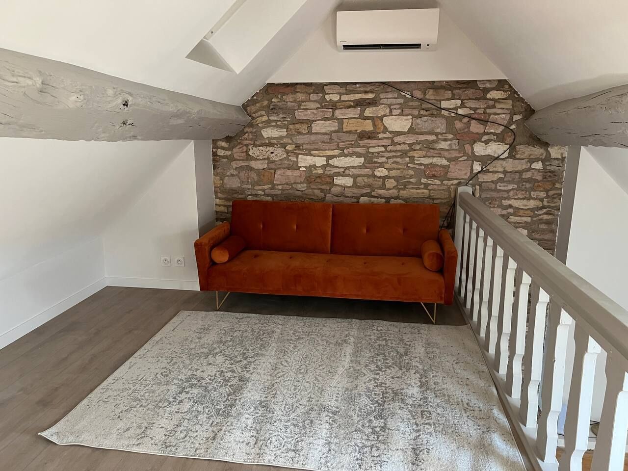 Un dormitorio tipo loft con pared de piedra, un sofá rojo, barandilla blanca y una alfombra estampada.