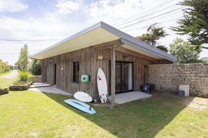 A wooden building with a covered porch, a stone wall, and a surfboard leaning against the wall. - Gîtes de Lézinadou - 8 personnes - Plomeur La Torche (plomeur)
