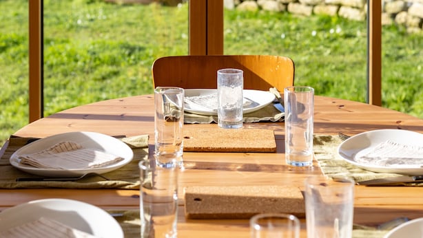 A wooden dining table set with plates, glasses, and cutlery, with a view of a stone wall and greenery outside.