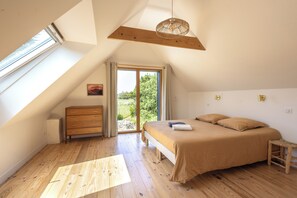 A bedroom with a sloped ceiling, a bed with a beige bedspread, a wooden dresser, and a view of a green landscape through a glass door. - Gîtes de Lézinadou - 6 personnes - Plomeur La Torche (plomeur)