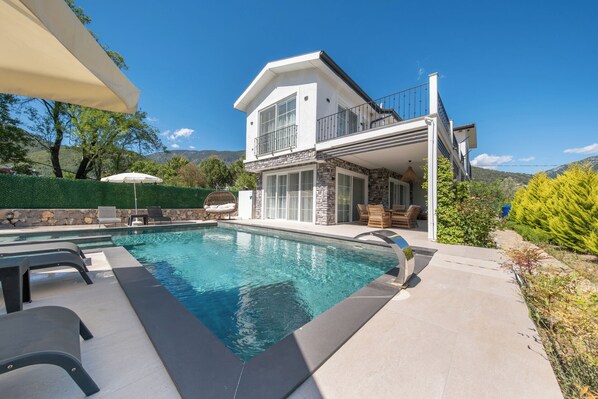 A modern two-story house with a swimming pool, outdoor furniture, and a stone facade.