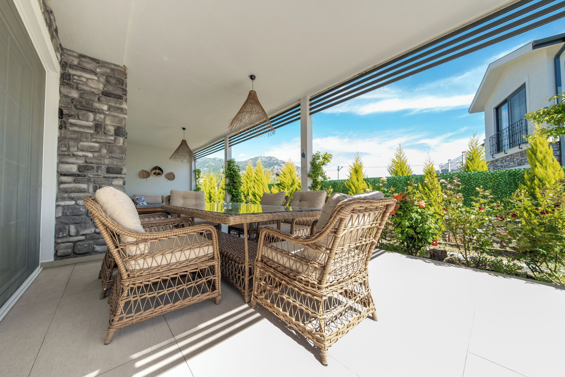 Une terrasse extérieure aménagée avec des meubles en osier, une table en verre et une vue sur un jardin arboré et fleuri.