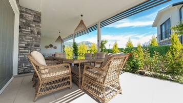 Une terrasse extérieure aménagée avec des meubles en osier, une table en verre et une vue sur un jardin arboré et fleuri.