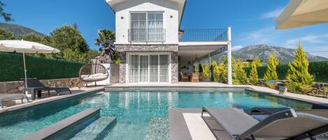 A two-story house with a swimming pool, outdoor furniture, and a mountain backdrop.
