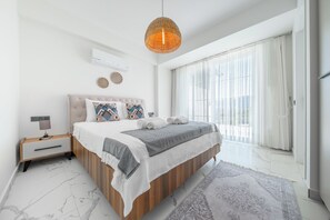 A modern bedroom with a large bed, a bedside table, a hanging light fixture, and a view of the outdoors through a large window.