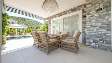 En terrasse med fletmøbler, en stenvæg og udsigt til et poolområde og grøn bevoksning.