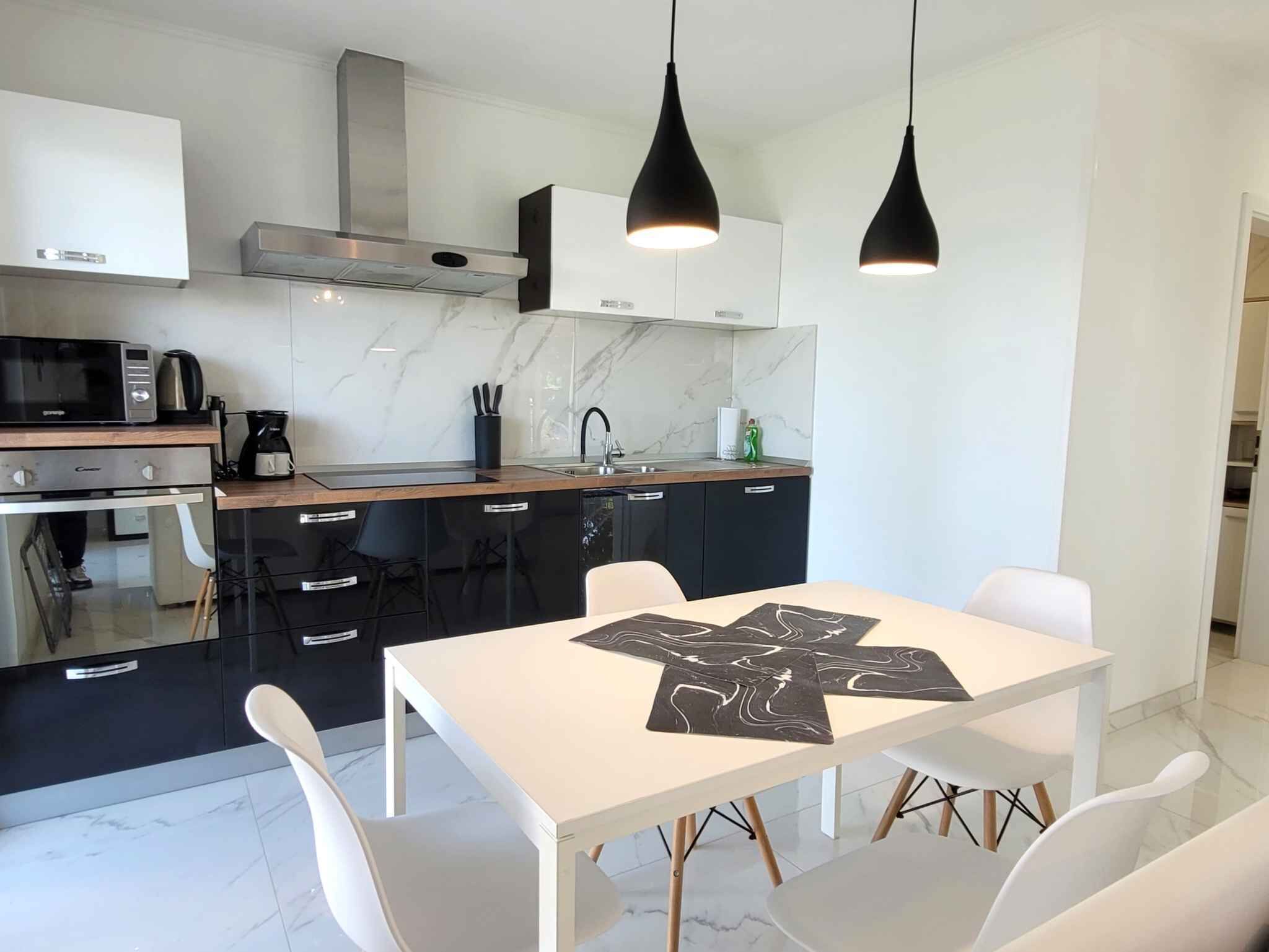 Una cocina moderna con encimera blanca, armarios negros y una campana extractora de acero inoxidable. Dos lámparas colgantes iluminan la isla de la cocina, que cuenta con una mesa blanca y caminos de mesa negros.