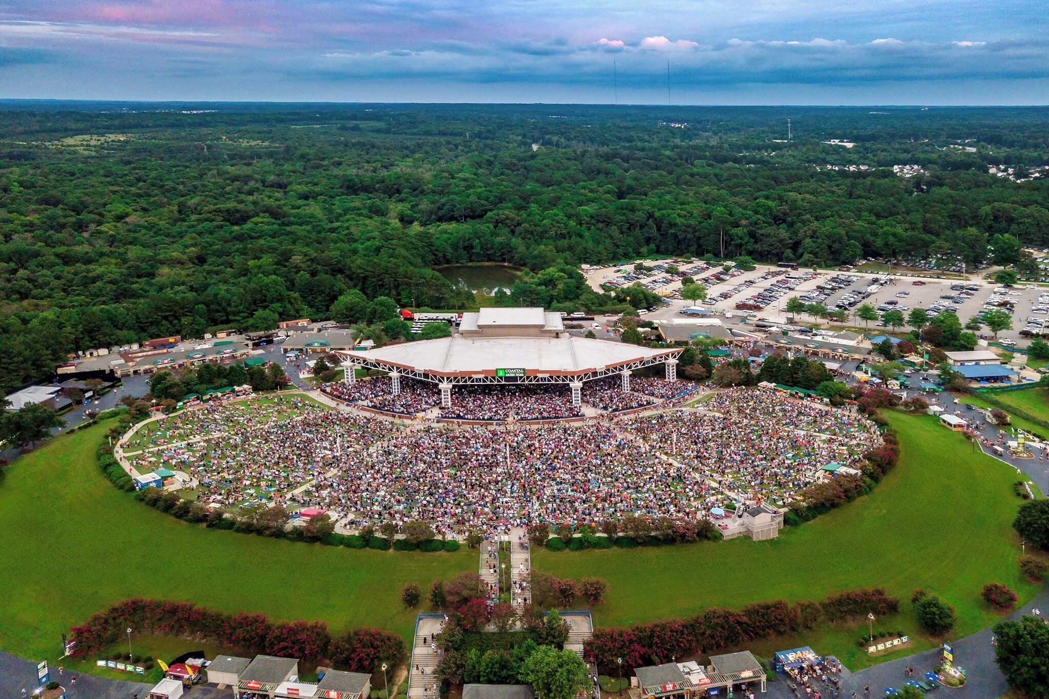 An aerial view of a stadium with a large crowd, surrounded by a parking lot and greenery.