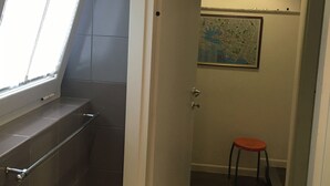 Un bagno con una finestra, un appendiabiti per asciugamani, una porta che dà su un'altra stanza e un piccolo sgabello.