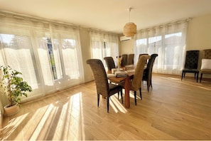 Un coin repas avec une table et des chaises en bois, de grandes fenêtres ornées de rideaux légers et un luminaire suspendu.