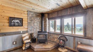 A cozy room with a stone fireplace, wooden walls, and a large window offering a view of the outdoors. - Peaceful Wyoming Cabin w/ Spacious Deck & Wet Bar! (Sundance)