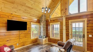 A log cabin interior with a stone fireplace, wooden walls, a flat-screen TV, a sofa, and a balcony with a view. - Peaceful Wyoming Cabin w/ Spacious Deck & Wet Bar! (Sundance)