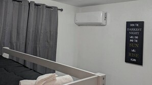 Ein schlicht eingerichtetes Schlafzimmer mit Etagenbett, grauen Vorhängen, einer Klimaanlage und einem motivierenden Zitat an der Wand.