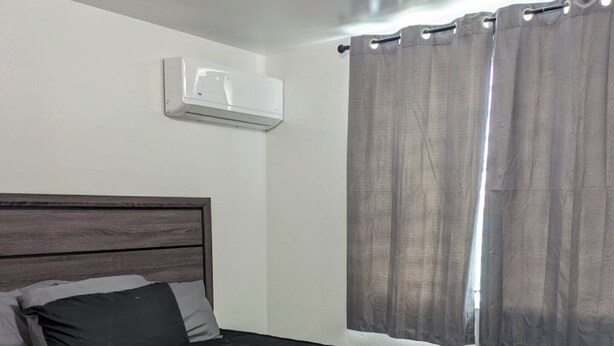 Ein Schlafzimmer mit einem dunkelbraunen Bett, einem Kopfteil, einer Wand-Klimaanlage und einem Fenster mit Vorhängen.