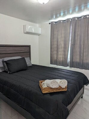 Une chambre à coucher avec un lit de couleur sombre, une tête de lit, un climatiseur fixé au mur et une fenêtre avec des rideaux.