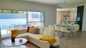 A modern living room with a white sofa, a round coffee table, and a dining area with a glass table and metal chairs. - Fantastic holiday home with heated pool (azeitão)