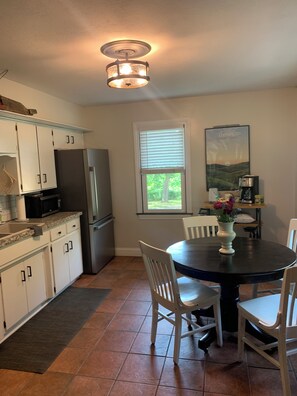 A kitchen with white cabinets, a black refrigerator, a dining table with white chairs, and a window with blinds.