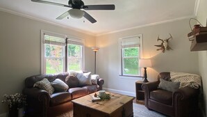 A living room with a brown leather sofa, a wooden coffee table, and a ceiling fan. There are two windows with blinds, a lamp, and a decorative skull on the wall. - Whitley Woods Cottage, secluded on 80 acres, Chadwick MO (Chadwick)