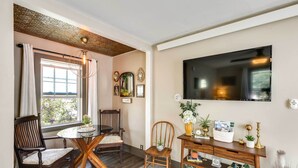 Un comedor con una mesa redonda y sillas, una consola de madera con objetos decorativos, un televisor montado en la pared y una ventana con cortinas.
