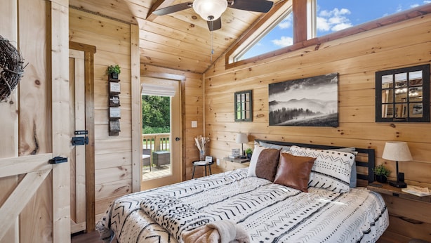 A wooden cabin bedroom with a bed, a ceiling fan, a window, and a painting on the wall.