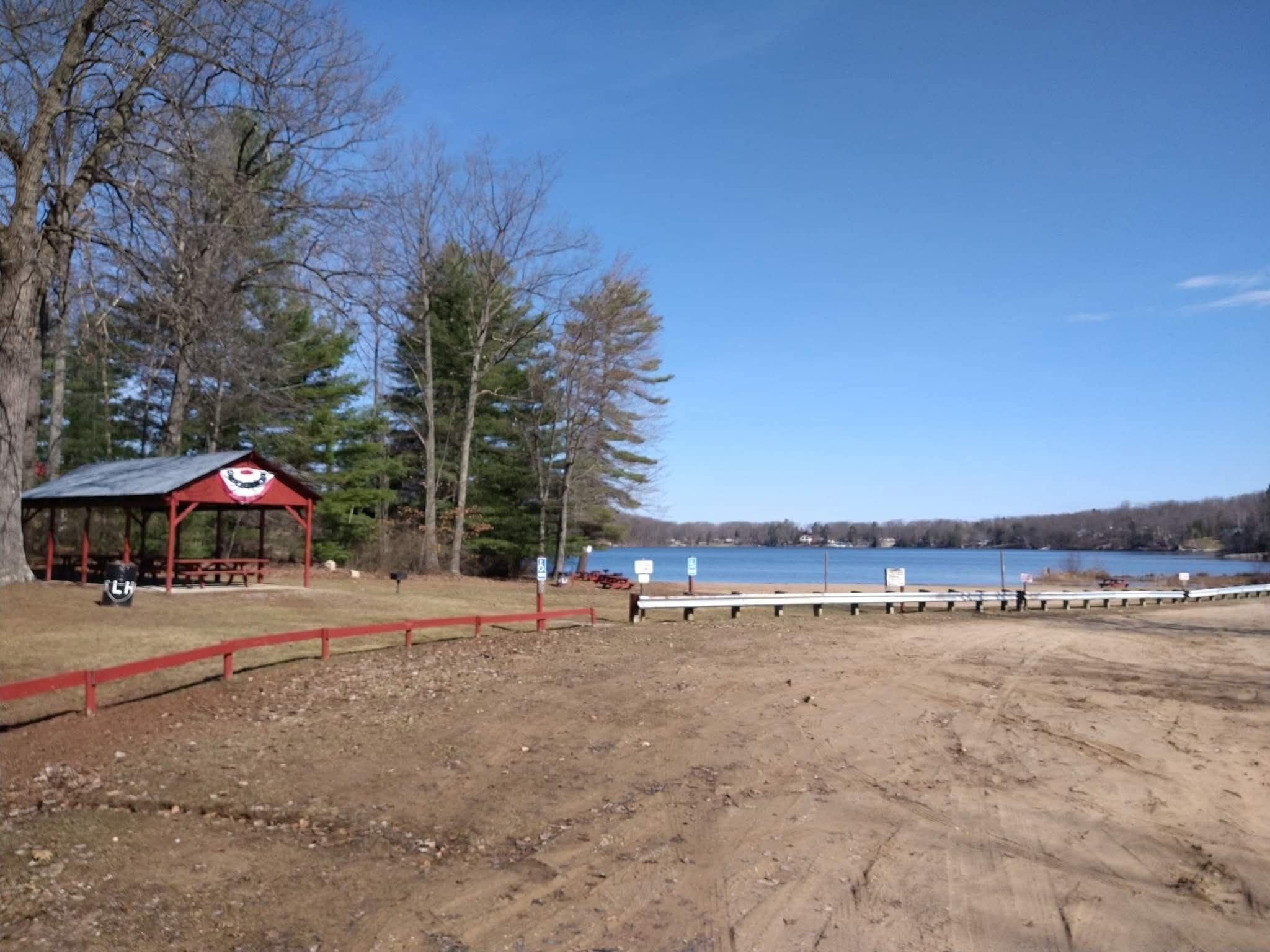 Un parc au bord du lac, avec un pavillon rouge, des tables de pique-nique et un sentier de terre qui mène à l’eau.