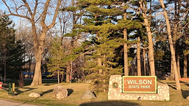 A sign for Wilson State Park with a stone wall and a grassy area.