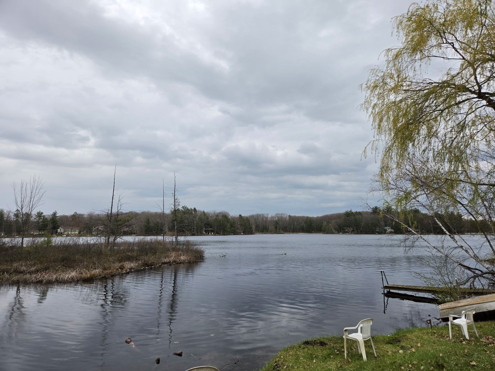 A calm lake with a dock and chairs, surrounded by trees and a cloudy sky.