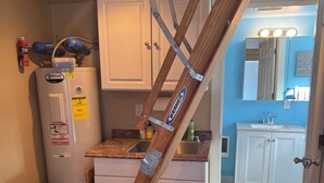 Una cocina con armarios blancos, un refrigerador de acero inoxidable, un bote de basura y una escalera de madera apoyada en la pared.