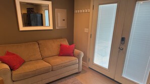 Un sofá color beige con cojines rojos, un espejo, una puerta con pomo y una ventana con persianas.