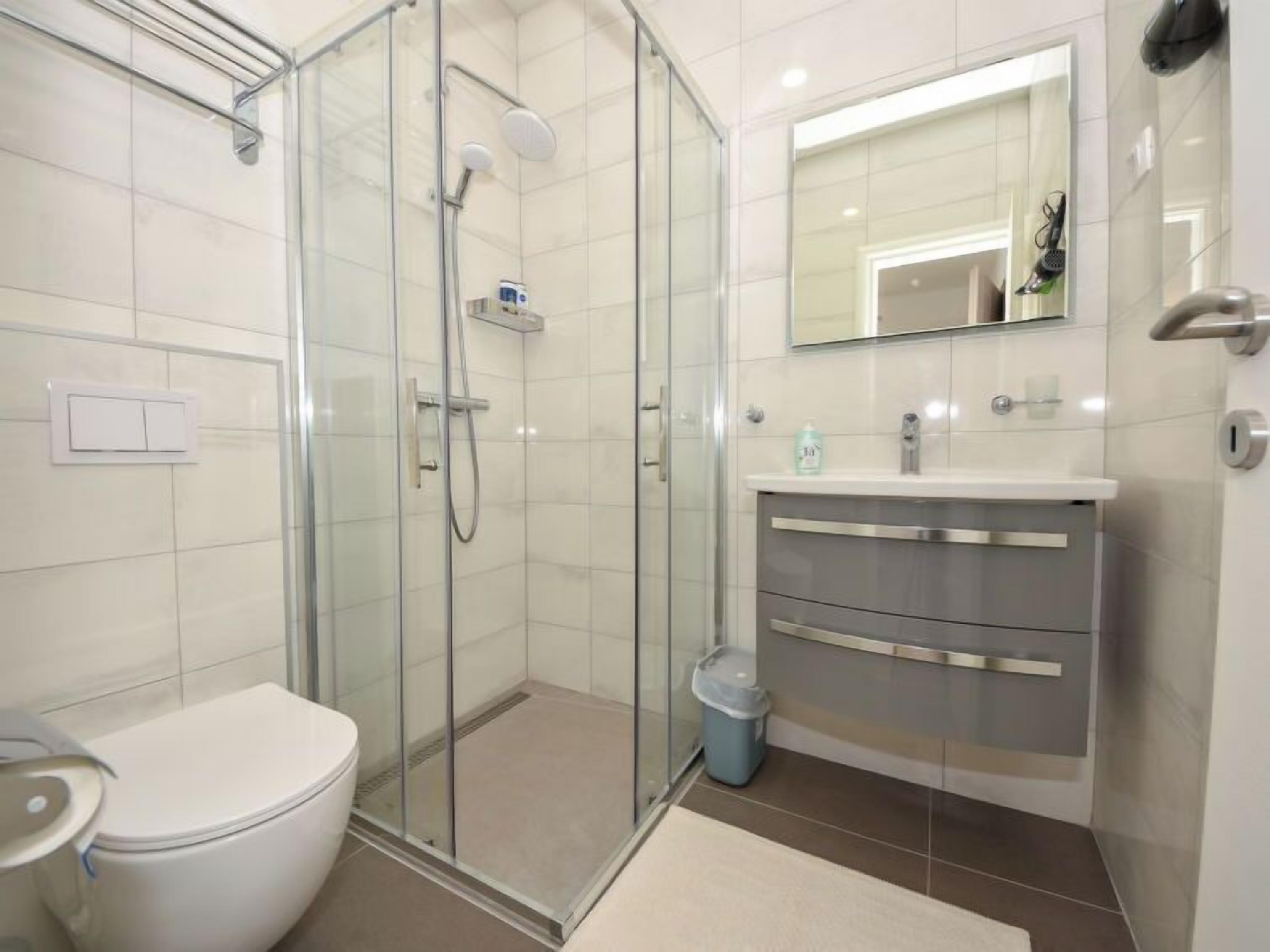 Un baño moderno con ducha envuelta en vidrio, inodoro blanco, tocador con espejo y un cesto de basura.
