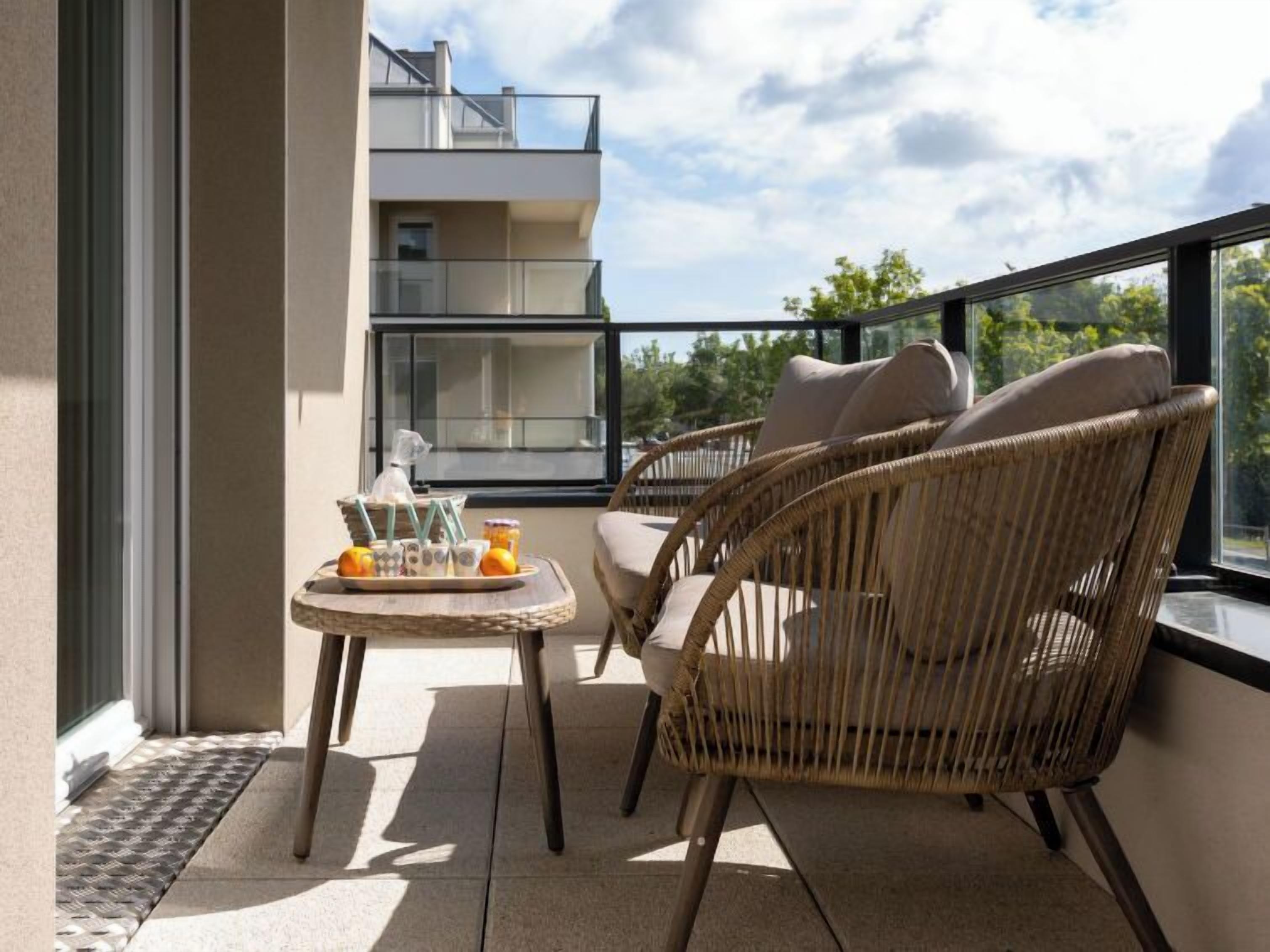 En balkong med flätade stolar och ett litet bord, med utsikt över en modern byggnad och grönområden.