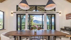A dining area with a large wooden table, white upholstered chairs, and a view of mountains through large windows. - Breathtaking Big Sky Luxury Retreat (Big Sky)